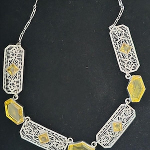 Op de afbeelding: Zilverkleurige filigraanhalsketting met gele edelsteenaccenten. De ketting heeft geometrische vormen en een fijne ketting. De edelstenen hebben verschillende vormen, waaronder vierkanten en zeshoeken. De ketting wordt tegen een donkere achtergrond getoond.