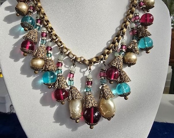 May include: A gold chain necklace with multiple strands of beads in shades of turquoise, pink, and white. The necklace features a variety of glass beads, including round, teardrop, and bell-shaped beads. The necklace is displayed on a white mannequin.