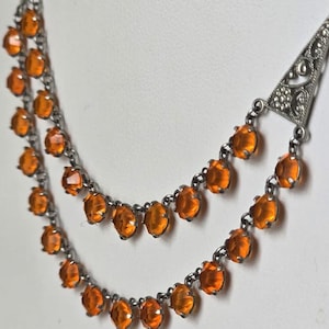 May include: A vintage silver-tone necklace with two strands of orange-colored gemstones. The necklace features a decorative triangular clasp at the top and is set against a white background. The gemstones are round and faceted.