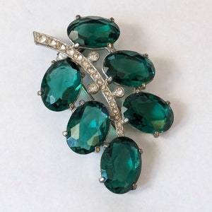 Könnte beinhalten: Eine silberfarbene Brosche mit einem Cluster aus ovalen, smaragdgrünen Strasssteinen. Die Strasssteine sind in silberfarbenem Metall gefasst und in Blattform angeordnet.