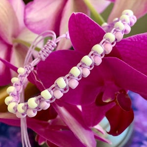 May include: A handmade bracelet featuring a light purple cord woven with alternating matte white and soft lavender beads. The design includes an adjustable sliding knot closure with two dangling cord ends, each finished with a single small bead.
