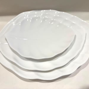 May include: Four white, leaf-shaped ceramic plates are stacked. The plates are of varying sizes, with a large plate at the base and a smaller one at the top. The plates have a wavy, textured edge.