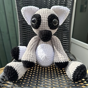 May include: A crocheted plush toy in the shape of a lemur. The lemur is gray with white fur on its belly and black accents around its eyes and on its tail. The lemur is sitting on a black wicker chair.