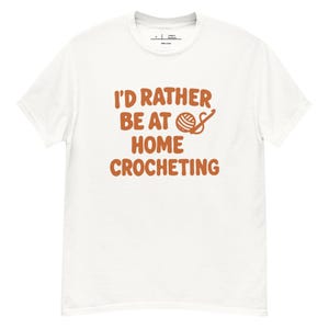 Peut inclure: T-shirt blanc avec l'inscription "I'D RATHER BE AT HOME CROCHETING" en orange. Le motif comprend une pelote de laine et un crochet. Le t-shirt a un col rond et des manches courtes, idéal pour un usage quotidien.