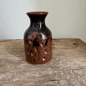 Könnte beinhalten: Eine kleine, braune und schwarze Keramikvase mit einer strukturierten Oberfläche. Die Vase hat einen runden Körper und einen schmalen Hals.