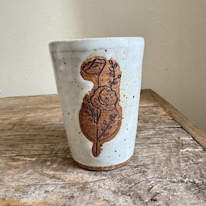 May include: White ceramic mug with a brown floral design. The mug has a speckled glaze and a brown rim.
