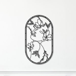 May include: Black metal wall art featuring a stylized line drawing of a woman's face surrounded by flowers and leaves. The art is oval shaped and has a simple, modern design. The text "Maya Metal Wall Art" is displayed below the art.