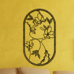 May include: Black metal wall art featuring a stylized outline of a woman's face surrounded by flowers and leaves. The art is mounted on a yellow wall above a brown couch.