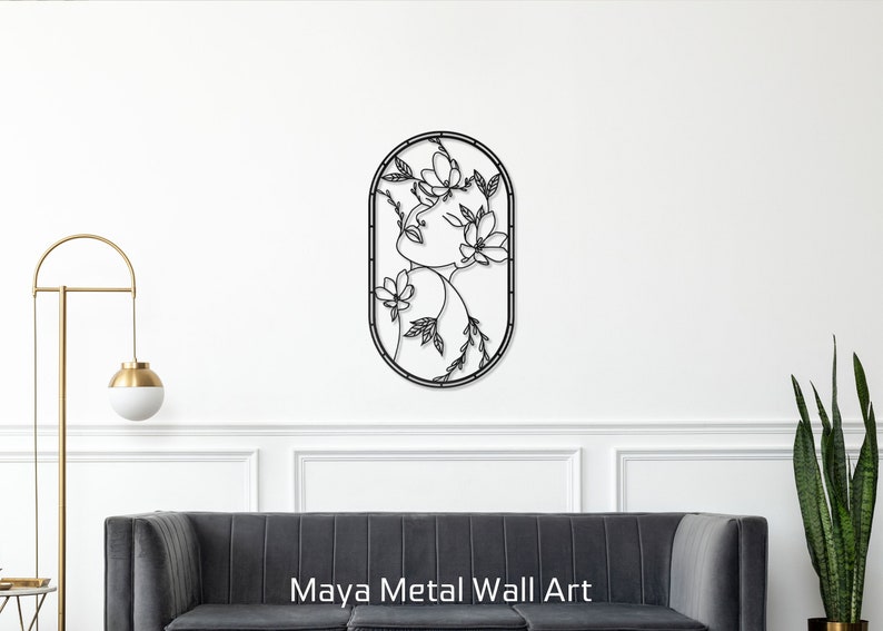 May include: Black metal wall art featuring a stylized line drawing of a woman's face surrounded by flowers and leaves. The art is oval shaped and has a simple, modern design. The text "Maya Metal Wall Art" is displayed below the art.