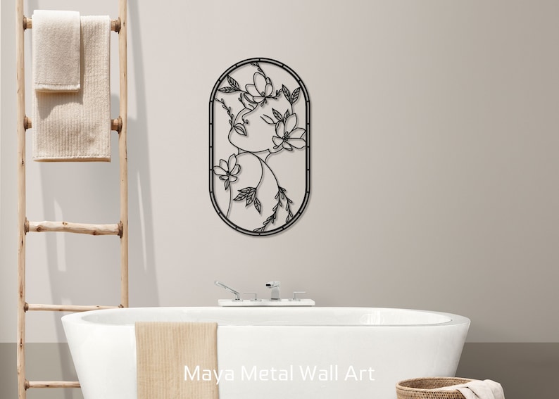 May include: Black metal wall art featuring a stylized line drawing of a woman's face with flowers in her hair. The art is in an oval frame and is hanging on a white wall above a bathtub.