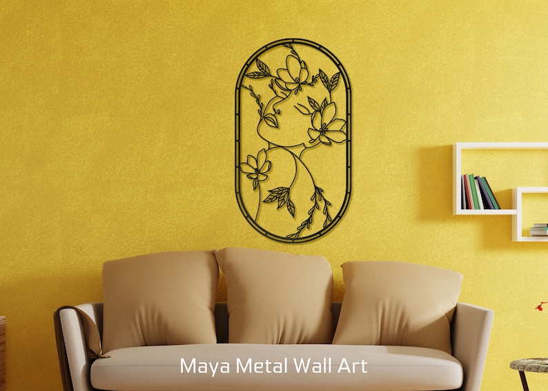 May include: Black metal wall art featuring a stylized outline of a woman's face surrounded by flowers and leaves. The art is mounted on a yellow wall above a brown couch.
