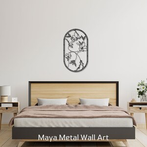 May include: Black metal wall art featuring a stylized floral design inside an oval frame. The art is hanging on a white wall above a wooden bed frame with a brown comforter.