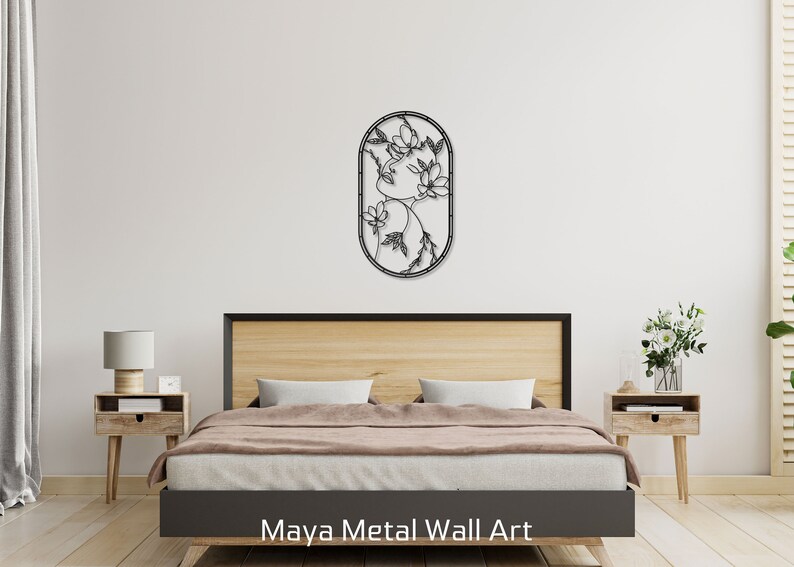 May include: Black metal wall art featuring a stylized floral design inside an oval frame. The art is hanging on a white wall above a wooden bed frame with a brown comforter.