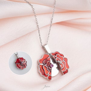 Puede incluir: Un collar de plata con un colgante de flor de esmalte rojo. El colgante es un medallón que se abre para revelar una pequeña piedra preciosa púrpura.