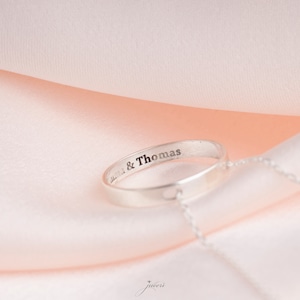 Puede incluir: Un anillo de plata con un corazón recortado y una cadena adjunta. El anillo está grabado con el texto "Anna & Thomas".