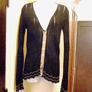 May include: A black long-sleeved cardigan featuring a deep V-neckline, a light beige button-down placket, and a subtle wavy pattern along the hem and cuffs. The knit fabric has a textured appearance with vertical detailing along the shoulders.