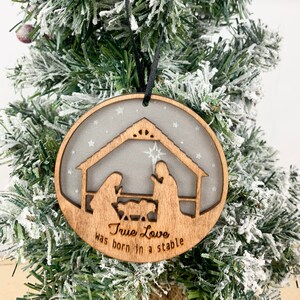 May include: A wooden Christmas ornament with a silhouette of the nativity scene. The ornament is decorated with white glitter and the text "True Love was born in a stable".