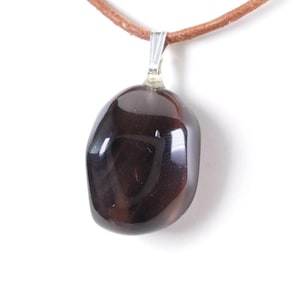 Puede incluir: Un colgante de piedra preciosa pulida de color marrón oscuro, con una fianza plateada, suspendido de un cordón de cuero marrón. La piedra tiene una forma lisa y redondeada y un acabado brillante, que refleja la luz. El collar se muestra sobre un fondo blanco.