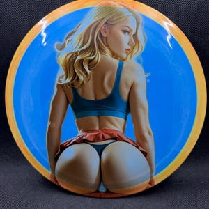 May include: A blue and orange disc golf disc featuring a woman with blonde hair, wearing a teal sports bra and a red skirt. The disc has a blue center with an orange and yellow outer ring.