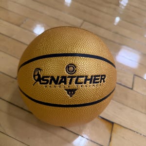 May include: A gold basketball with black accent lines and the word "SNATCHER" printed in black. The ball also has the words "HANDLE TRAINING" printed in black. The basketball is sitting on a wooden floor.