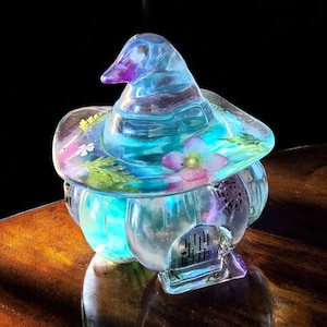 May include: A blue glass witch hat with a small house inside. The hat is decorated with dried flowers and greenery.
