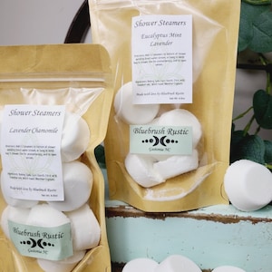 May include: Two kraft paper pouches filled with white shower steamers. One pouch is labeled "Lavender Chamomile," the other "Eucalyptus Mint Lavender." The labels include instructions and ingredients. The brand name "Bluebrush Rustic" is also visible.