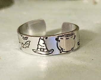 May include: A silver adjustable ring with a Halloween theme. The ring features a witch's hat, a cauldron, and bats.