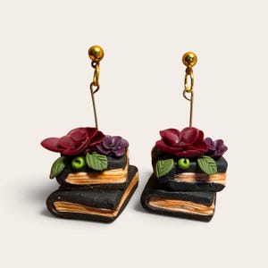 May include: A pair of dangle earrings featuring miniature stacks of two black books with tan pages. Each stack is topped with sculpted dark red and purple flowers and green leaves. The earrings hang from gold-toned ball stud posts.