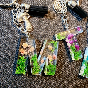 Puede incluir: Llaveros con letras de resina transparente, rellenas de flores secas y vegetación. La letra "M" presenta flores rosas y musgo verde. Los dijes plateados de hongos y loto cuelgan de los llaveros, junto con borlas negras.