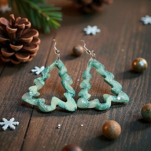 May include: A pair of Christmas tree-shaped earrings in shades of green with silver accents. The earrings are displayed on a wooden surface with pine cones, evergreen sprigs, and decorative elements.