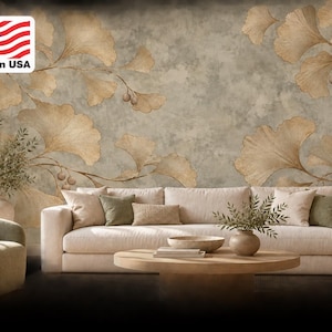 Könnte beinhalten: Ein Wandbild mit großen, beigen Ginkgoblättern auf einem strukturierten, fleckigen grauen Hintergrund. Das Wandbild befindet sich hinter einem beigen Sofa, einem runden Couchtisch aus Holz, einem grünen Sessel und einer Lampe. Der Text Made in USA ist sichtbar.