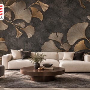 Könnte beinhalten: Ein Wandbild mit großen, goldenen Ginkgoblättern vor einem dunklen, strukturierten anthrazitfarbenen Hintergrund. Das Wandbild befindet sich hinter einem cremefarbenen Sofa, einem runden Couchtisch aus Holz und einem dunkelgrauen Sessel.