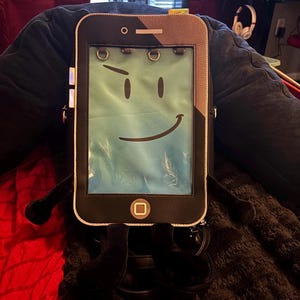 Könnte beinhalten: Ein schwarzer Rucksack in Form eines Smartphones mit einem durchsichtigen Sichtfenster auf der Vorderseite, das ein Cartoon-Gesicht mit hochgezogener Augenbraue und Lächeln zeigt. Der Rucksack hat einen quadratischen Home-Button und schwarze Plüschbeine.