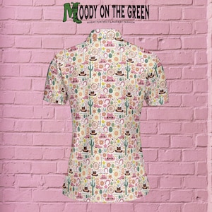 May include: A white short-sleeved button-down shirt with a colorful pattern of cowboy boots, hats, cacti, horseshoes, and wagon wheels. The text "MOODY ON THE GREEN" is printed on the back of the shirt.