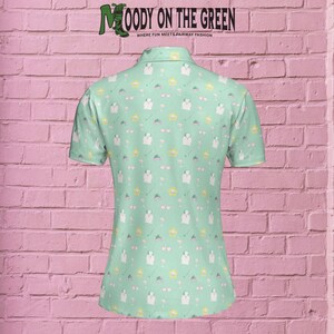 May include: A light green golf shirt with a white collar and short sleeves. The shirt features a repeating pattern of golf clubs, sunglasses, flowers, and hearts. The text "MOODY ON THE GREEN" is printed on the back of the shirt.