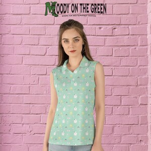 May include: A sleeveless green polo shirt with a white and yellow pattern. The shirt has a collared neckline and a button closure. The text "MOODY ON THE GREEN" is printed on the shirt. The text "WHERE FUN MEETS FAIRWAY FASHION" is printed below the main text.