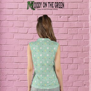 May include: A sleeveless, light green golf shirt with a white collar. The shirt has a repeating pattern of white golf carts, golf clubs, and golf balls. The text "MOODY ON THE GREEN" is printed on the back of the shirt.