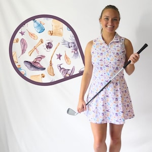 May include: A sleeveless golf dress with a collared neckline, featuring a light pink background and a repeating pattern of witch-themed illustrations. The dress is paired with a golf club. A circular graphic displays similar illustrations.