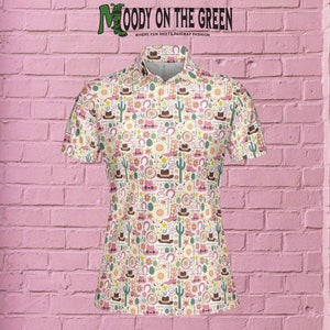 May include: A white short-sleeved golf shirt with a colorful pattern of western-themed icons, including cacti, horseshoes, cowboy hats, and boots. The shirt has a pink collar and a small logo that reads "MOODY ON THE GREEN, WHERE FUN MEETS FAIRWAY FASHION".