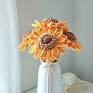 May include: A bouquet of crocheted sunflowers in a white vase. The sunflowers have yellow petals and brown centers. The vase is tied with a cream-colored string. The arrangement sits on a white surface.