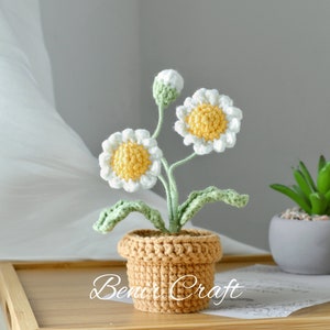 May include: A crocheted daisy flower plant in a brown pot. The plant has two white flowers with yellow centers and green leaves. The pot is sitting on a wooden tray. The text "Benir Craft" is visible on the tray.