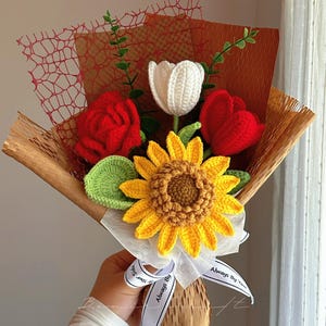 Puede incluir: Un ramo de flores de ganchillo con un girasol amarillo, un tulip&aacute;n blanco, una rosa roja y un tulip&aacute;n rojo con hojas verdes. El arreglo est&aacute; envuelto en papel marr&oacute;n y malla roja, atado con una cinta blanca con el texto Always By Your Side.
