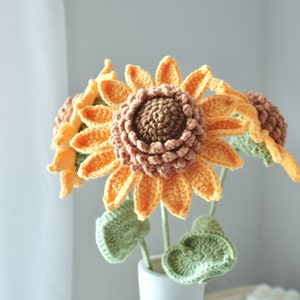 May include: A bouquet of handmade crochet sunflowers in a white vase. The sunflowers have orange petals, brown centers, and green leaves. The vase is tied with a natural twine. The flowers are a charming home decor item.