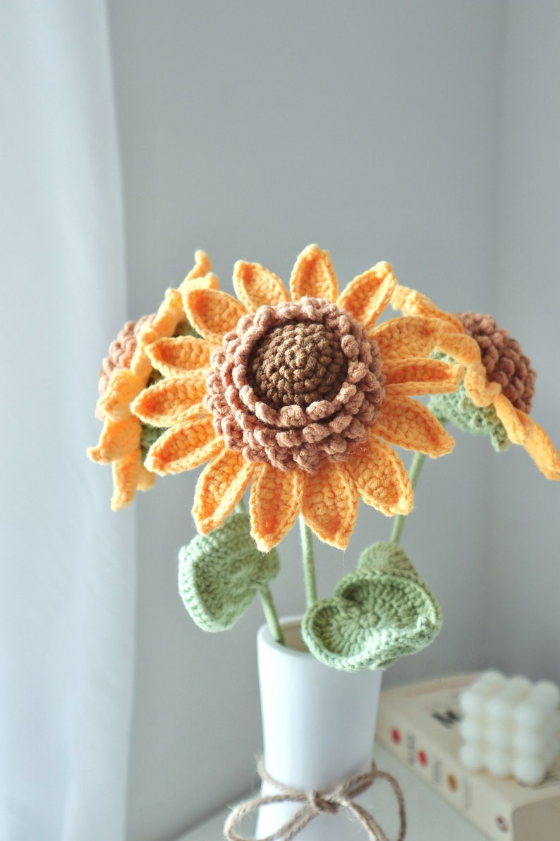 May include: A bouquet of handmade crochet sunflowers in a white vase. The sunflowers have orange petals, brown centers, and green leaves. The vase is tied with a natural twine. The flowers are a charming home decor item.