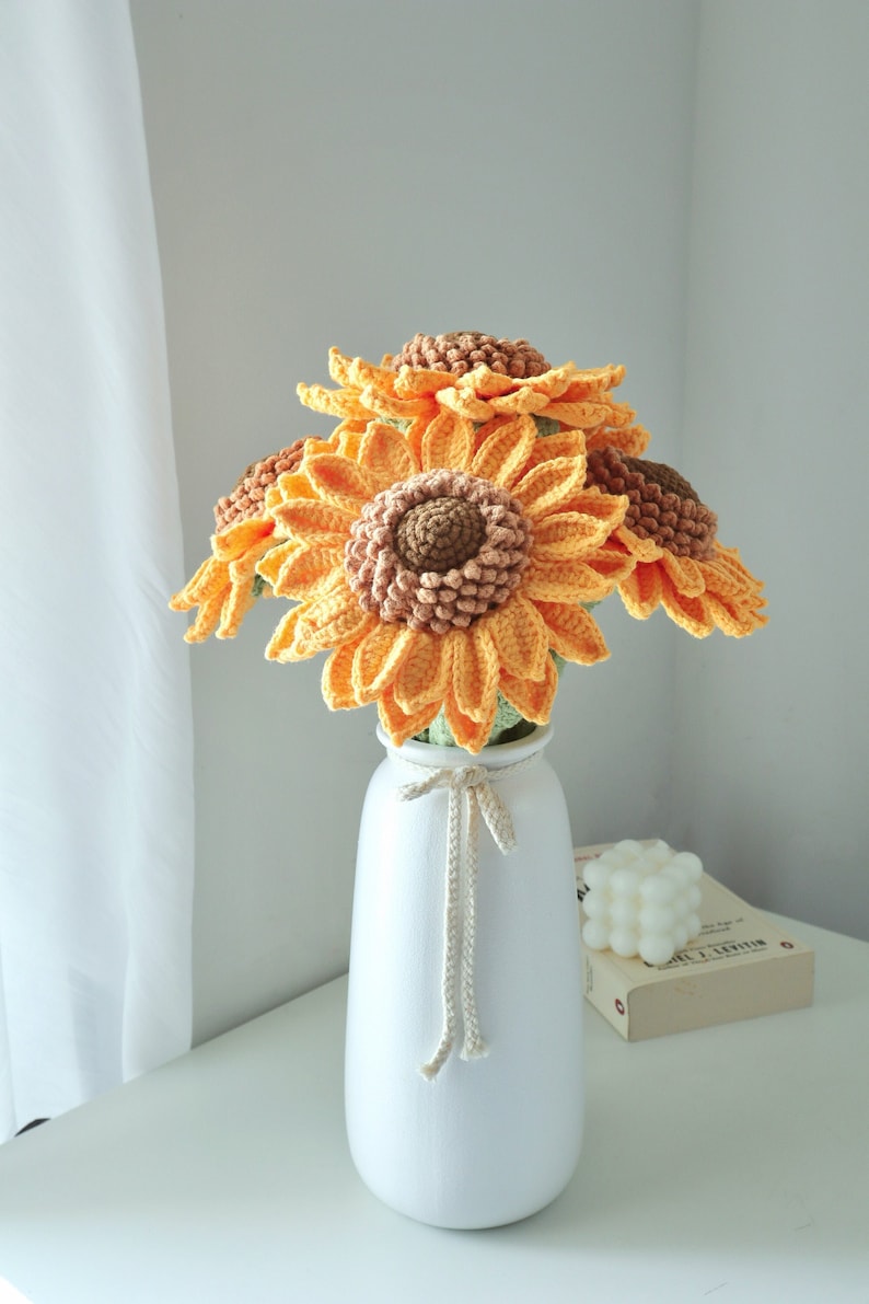May include: A bouquet of crocheted sunflowers in a white vase. The sunflowers have yellow petals and brown centers. The vase is tied with a cream-colored string. The arrangement sits on a white surface.