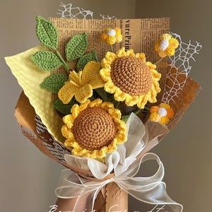 Puede incluir: Un ramo de flores de ganchillo con dos girasoles grandes de centros marrones y p&eacute;talos amarillos, un lirio amarillo y flores amarillas m&aacute;s peque&ntilde;as con hojas verdes. El ramo est&aacute; envuelto en papel marr&oacute;n, papel de seda amarillo y encaje blanco, atado con una cinta blanca.