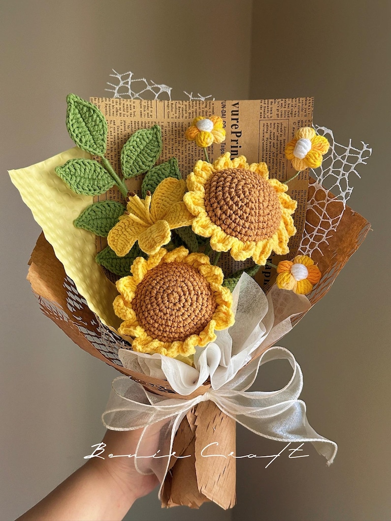 Puede incluir: Un ramo de flores de ganchillo con dos girasoles grandes de centros marrones y p&eacute;talos amarillos, un lirio amarillo y flores amarillas m&aacute;s peque&ntilde;as con hojas verdes. El ramo est&aacute; envuelto en papel marr&oacute;n, papel de seda amarillo y encaje blanco, atado con una cinta blanca.