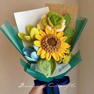 May include: A handmade crochet bouquet featuring a large yellow sunflower, blue and green flowers, and green leaves. The bouquet is wrapped in teal paper and tied with a navy blue ribbon. The bouquet is a unique, handcrafted gift.