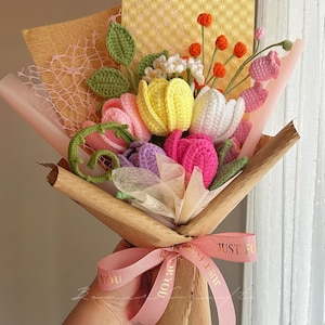 Puede incluir: Un ramo de flores de ganchillo hecho a mano con tulipanes amarillos, blancos, rosas y morados, peque&ntilde;as flores blancas y bayas naranjas. El arreglo incluye hojas verdes y est&aacute; envuelto en papel marr&oacute;n con una cinta rosa impresa con el texto JUST FOR YOU.