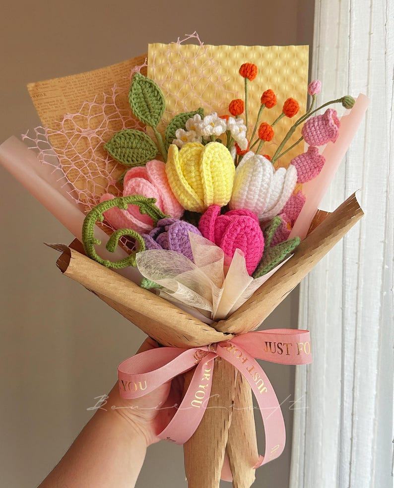 Puede incluir: Un ramo de flores de ganchillo hecho a mano con tulipanes amarillos, blancos, rosas y morados, peque&ntilde;as flores blancas y bayas naranjas. El arreglo incluye hojas verdes y est&aacute; envuelto en papel marr&oacute;n con una cinta rosa impresa con el texto JUST FOR YOU.